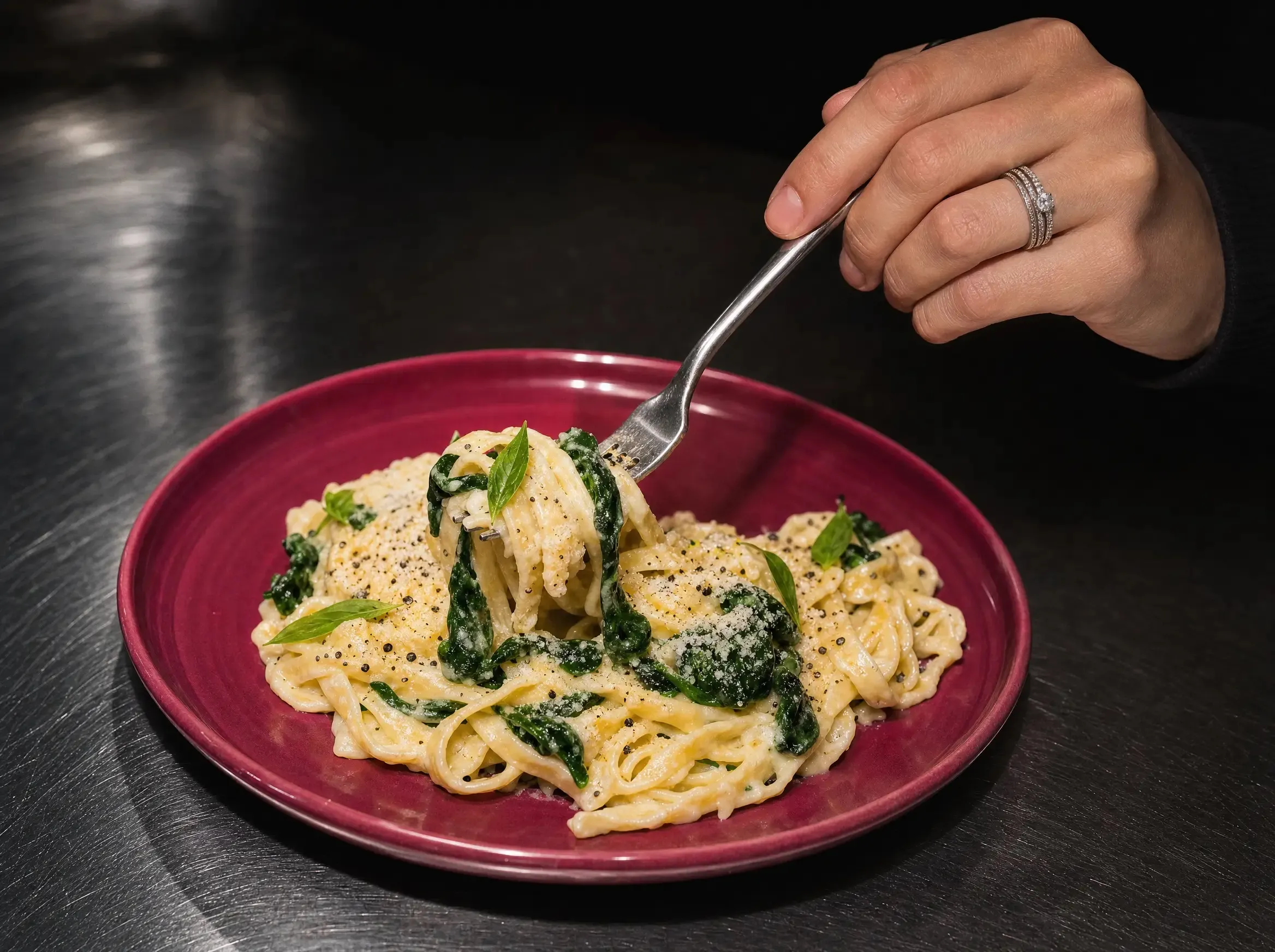 Eine Hand hebt mit der Gabel Tagliatelle mit Spinat und cremiger Zitronen-Ricotta-Sauce vom beerenfarbenen Teller.