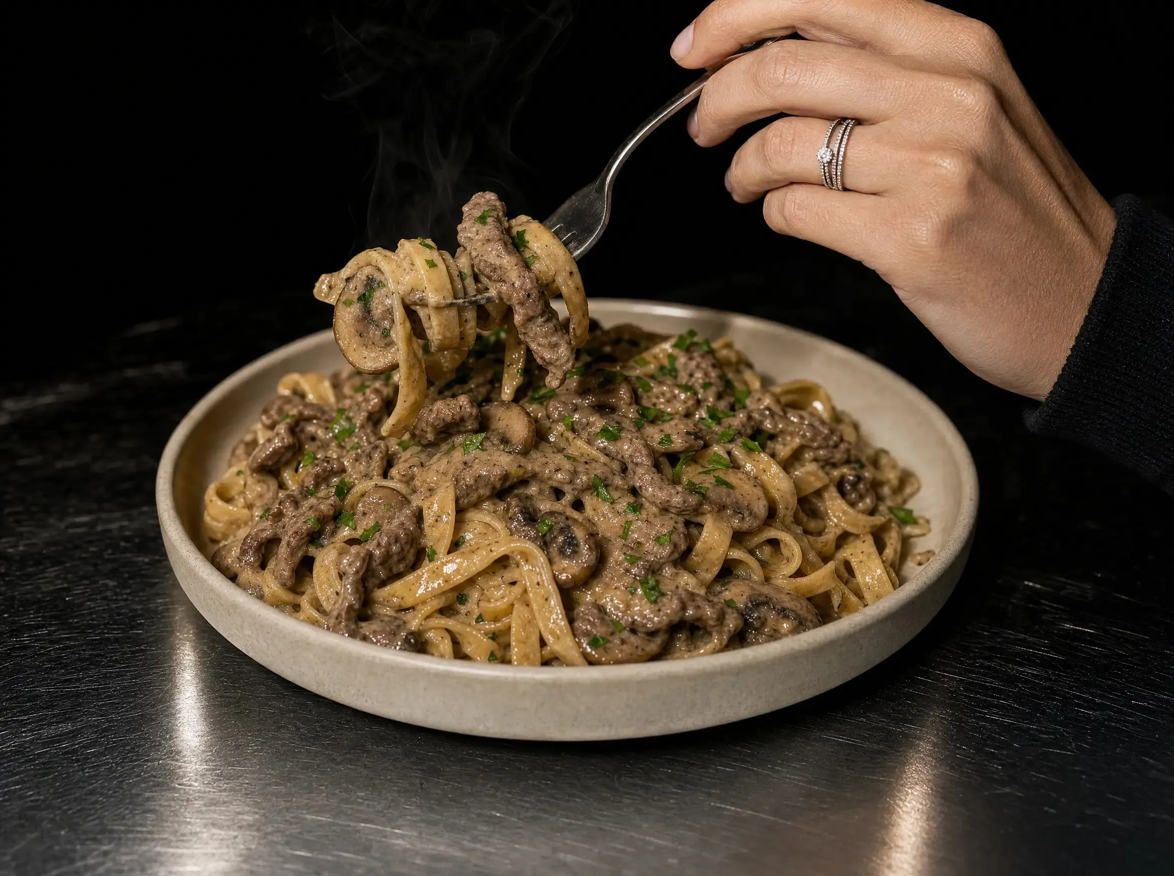 Eine Hand hebt mit der Gabel Tagliatelle, Rinderhack und Pilzscheiben aus der vollen Schüssel Beef Stroganoff, deutlich sichtbarer Dampf.