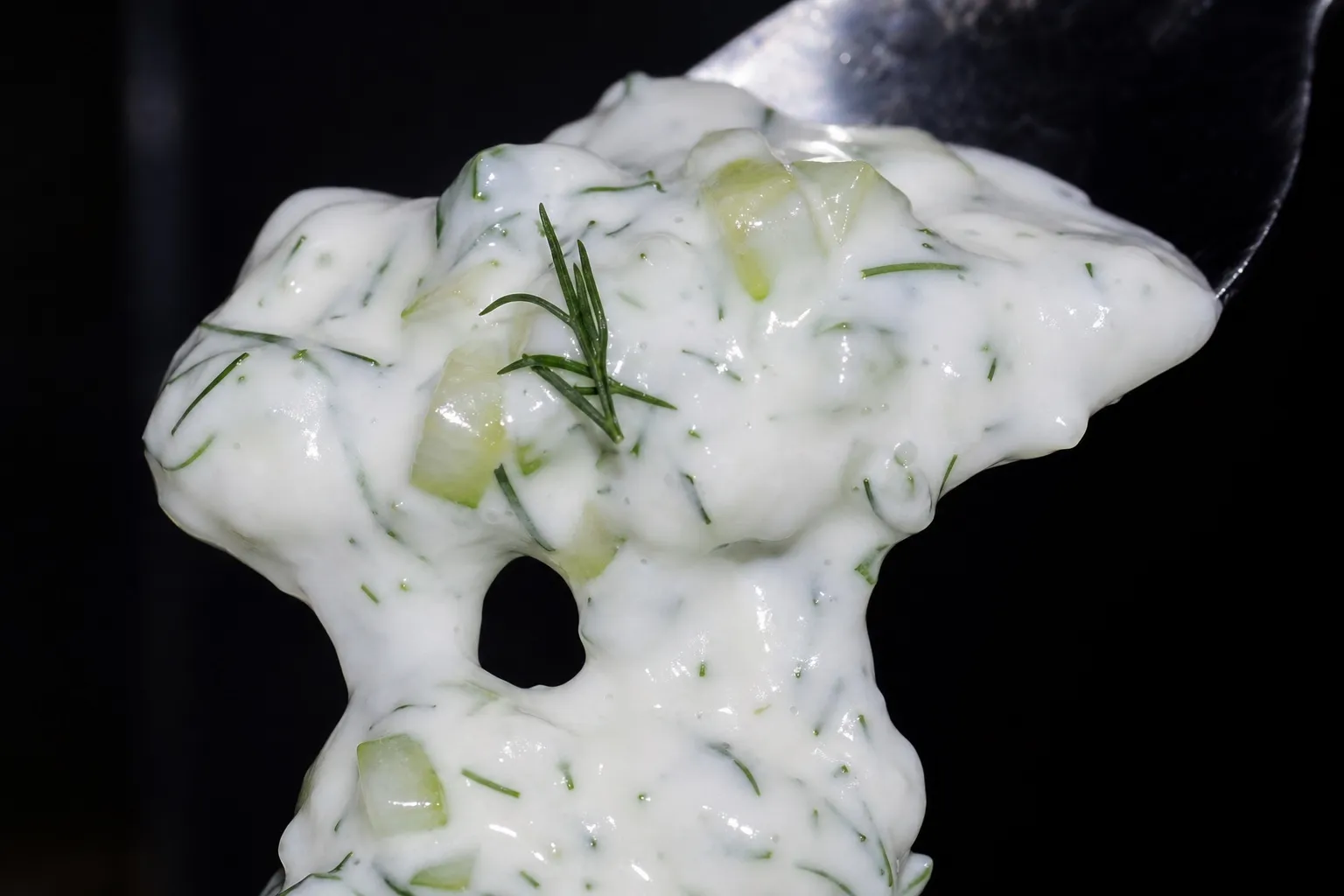 Nahaufnahme von cremigem Tzatziki mit Gurkenstücken und Dill, das an einem Löffel haftet und in dicken Strängen herunterläuft.