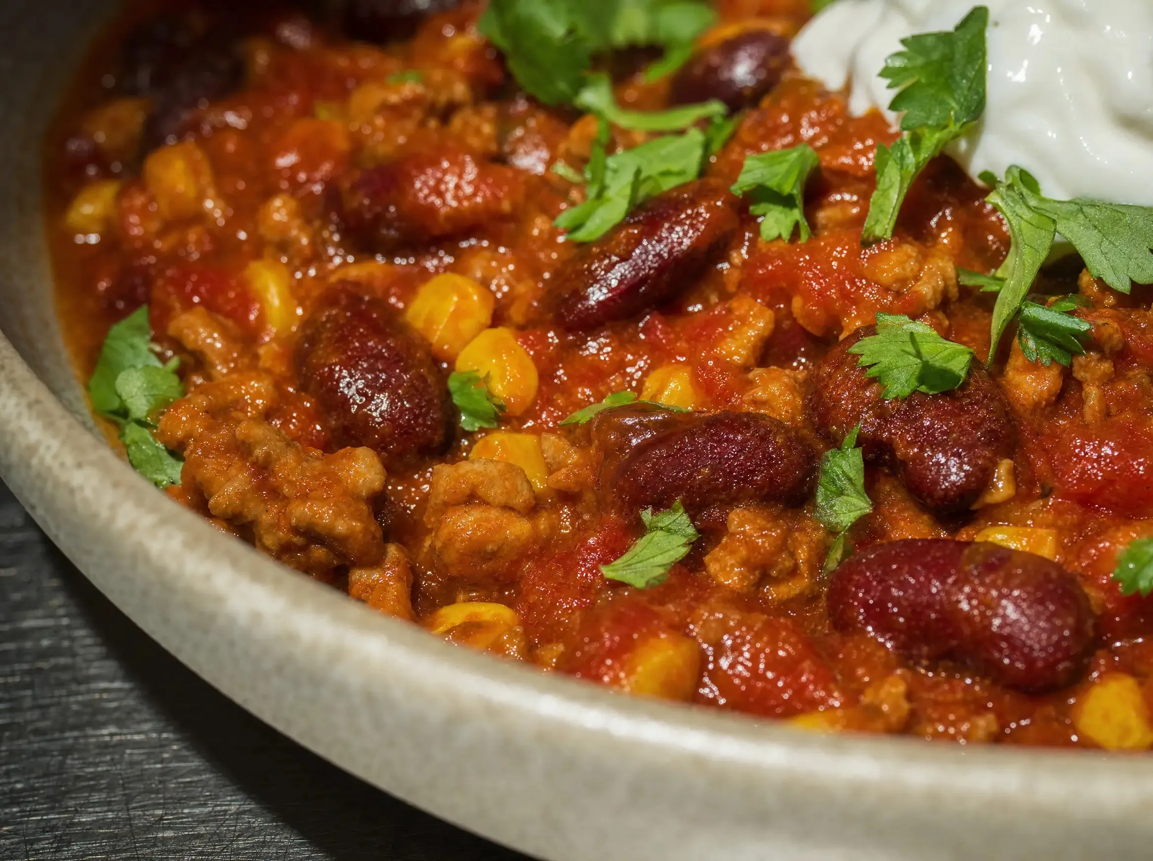 Nahaufnahme der Chili-Oberfläche mit Kidneybohnen, Mais und Sojaschnetzel in glänzender Sauce, Korianderblätter und ein Klecks Joghurt am Bildrand.