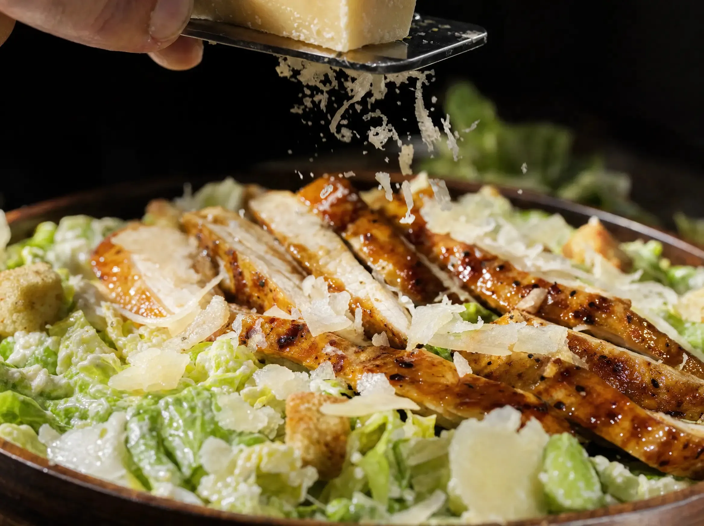Frisch gehobelter Parmesan fällt von einer Reibe über den fertigen High Protein Caesar Salad mit Hähnchenstreifen und Croutons.