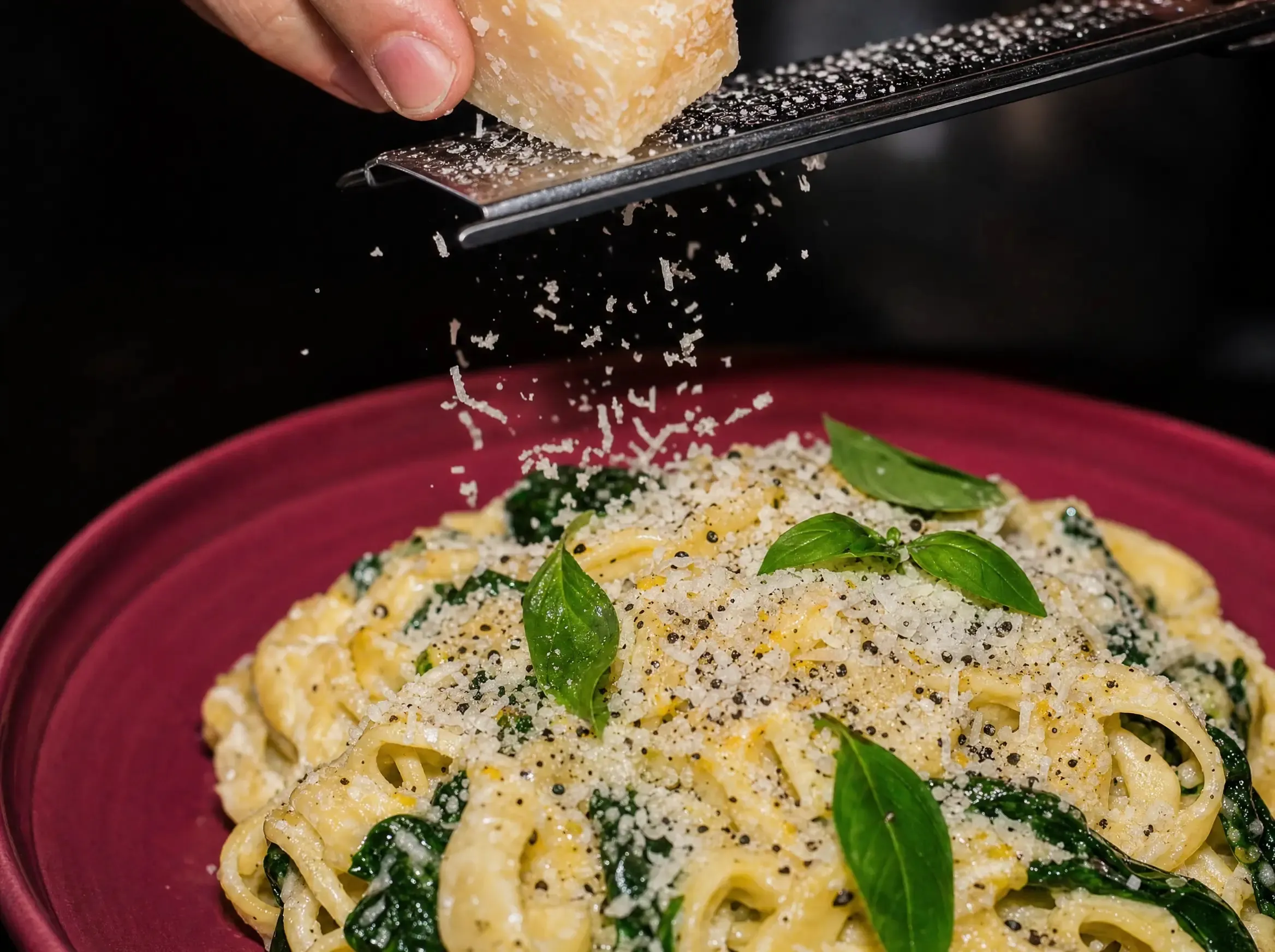Frisch geriebener Parmesan fällt von einer Reibe über die fertige Zitronen-Ricotta-Pasta auf dem beerenfarbenen Teller.