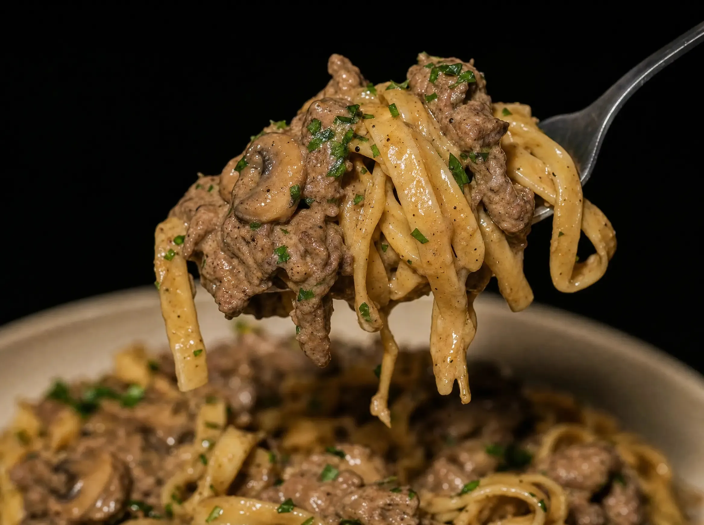 Makroaufnahme einer Gabel mit um sie gedrehten Tagliatelle, Rinderhack und Pilzen, cremige Sauce tropft zurück in die Schüssel.