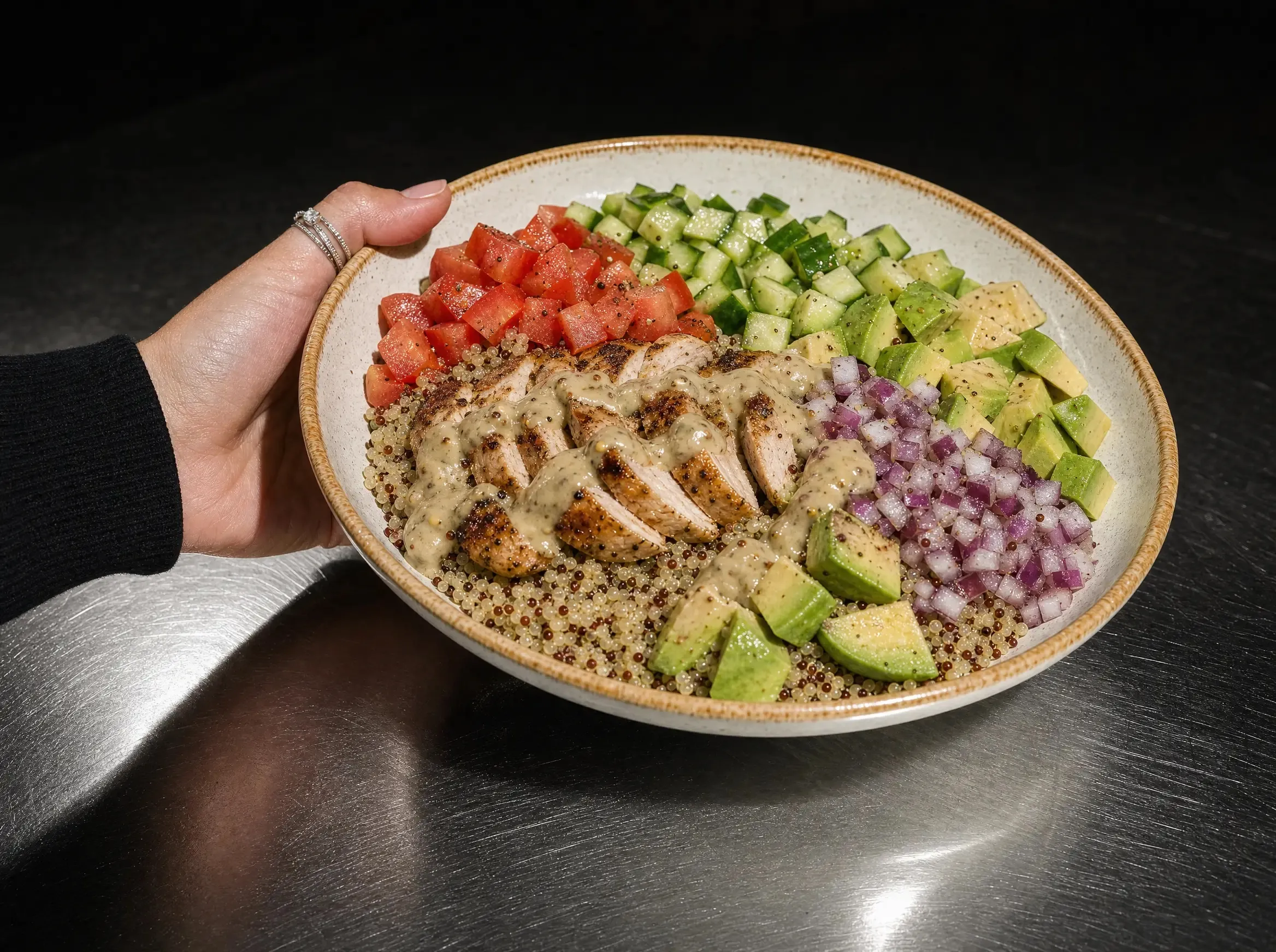 Eine Hand hält die Quinoa Bowl leicht zur Kamera geneigt über der dunklen Edelstahl-Arbeitsplatte.