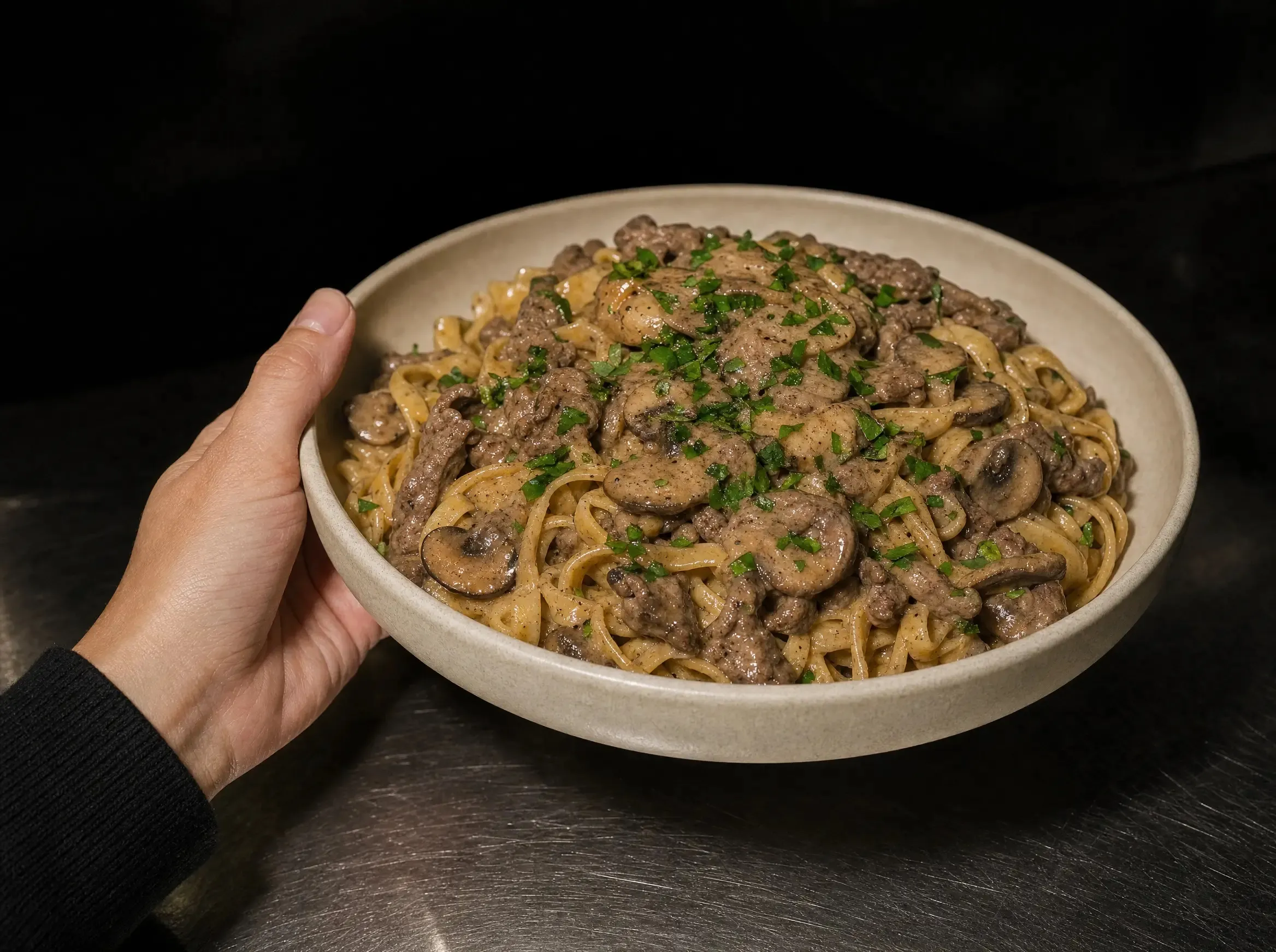 Eine Schüssel Beef Stroganoff mit Nudeln, Pilzen und Petersilie wird in einer Hand über der dunklen Edelstahl-Arbeitsplatte gehalten.