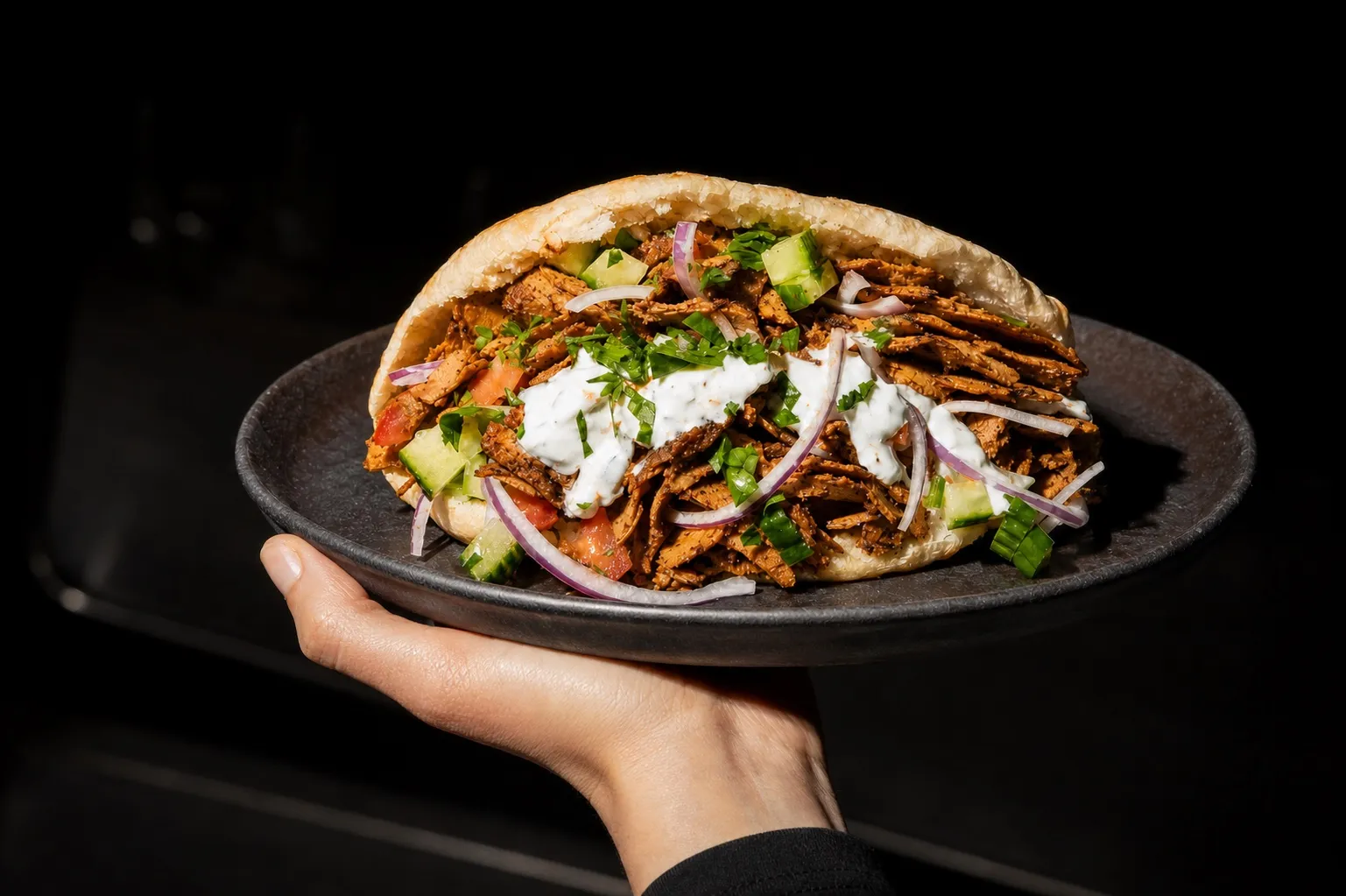 Frauenhand hält einen dunklen Keramikteller mit Tofu Döner im Pita-Brot, knusprige Räuchertofu-Streifen und frisches Gemüse sichtbar.