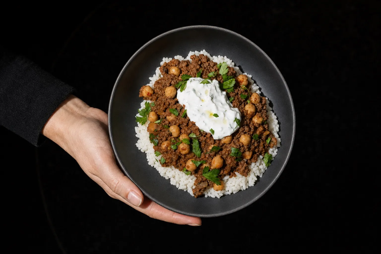 Frauenhand hält eine dunkle Keramikschüssel mit Hackfleisch-Kichererbsen-Pfanne und Joghurt-Topping.