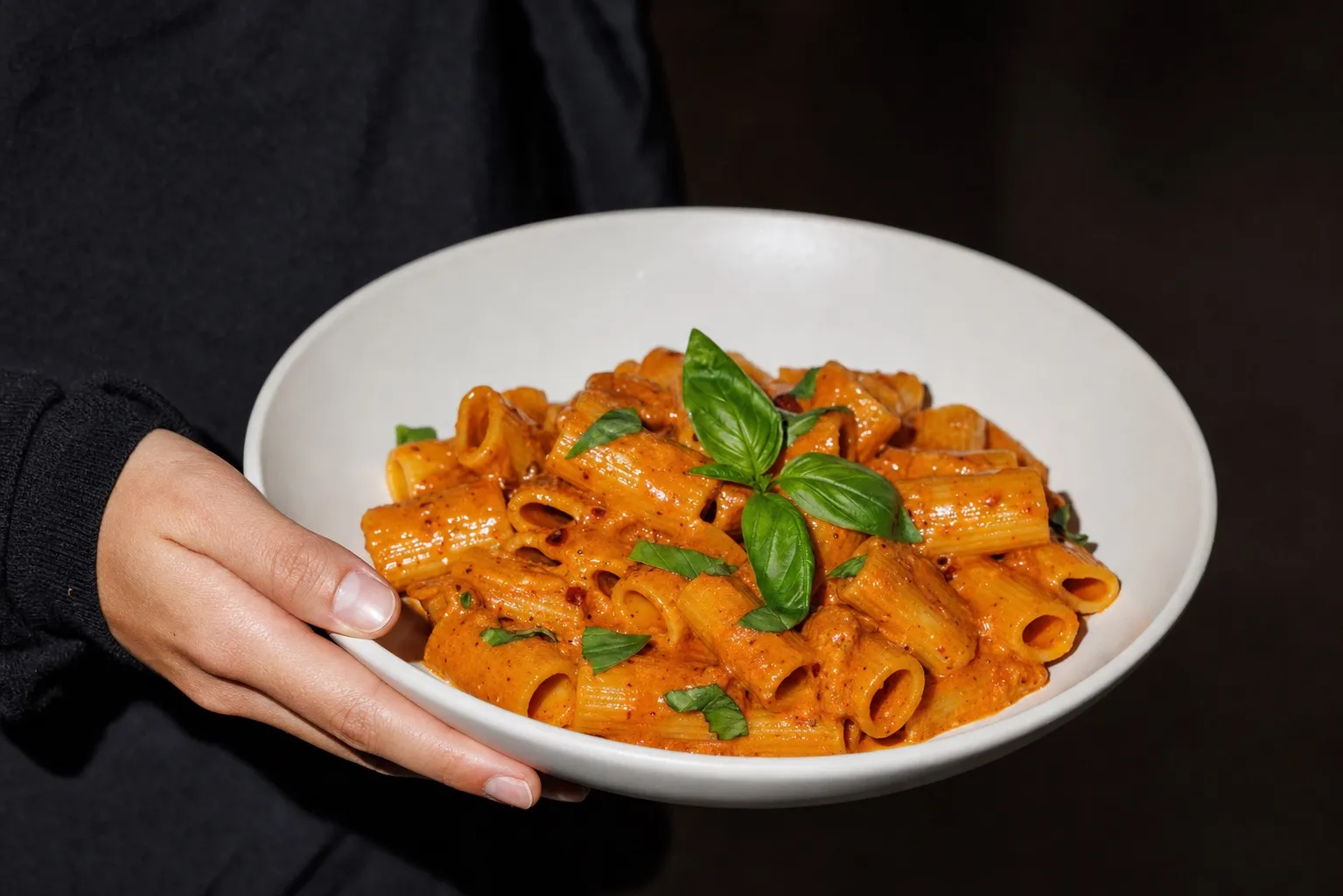 Frauenhand hält eine weiße Schale mit Rigatoni in cremiger Pasta alla Vodka, mit Basilikum garniert, leicht zur Kamera geneigt.