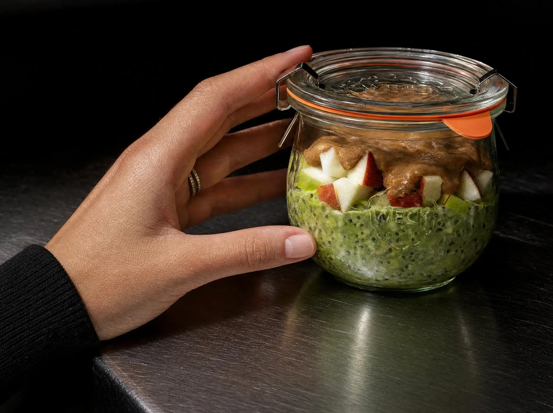 Ein WECK-Glas mit Matcha Overnight Oats, Apfelwürfeln und Nussmus wird in einer Hand auf der dunklen Edelstahl-Arbeitsplatte gehalten.