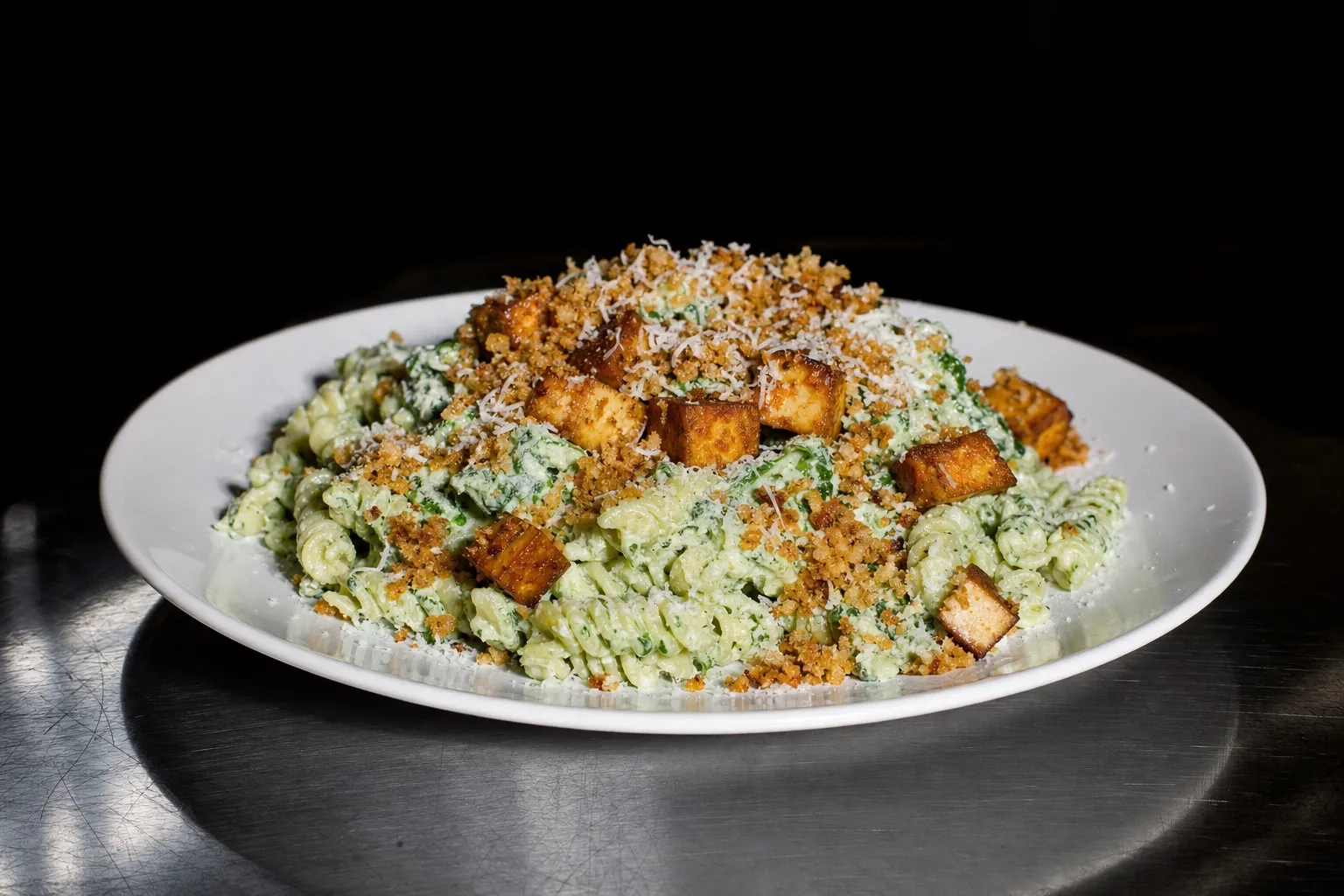 Teller mit Spinat-Pasta, dicht belegt mit Räuchertofu, Panko und Parmesan, vor dunklem Hintergrund.