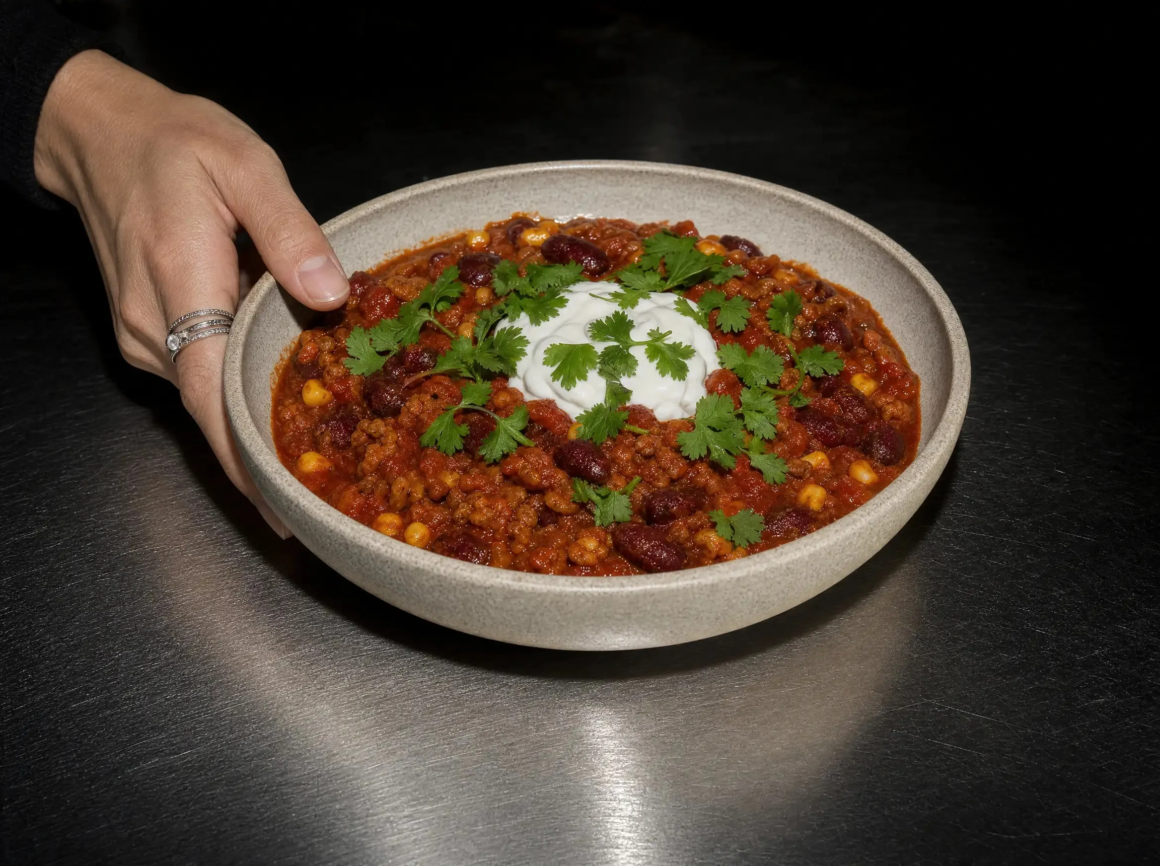 Eine Hand fasst eine Schale Chili sin Carne mit Joghurt und Koriander am Rand, die Schale steht auf einer dunklen Edelstahlfläche.