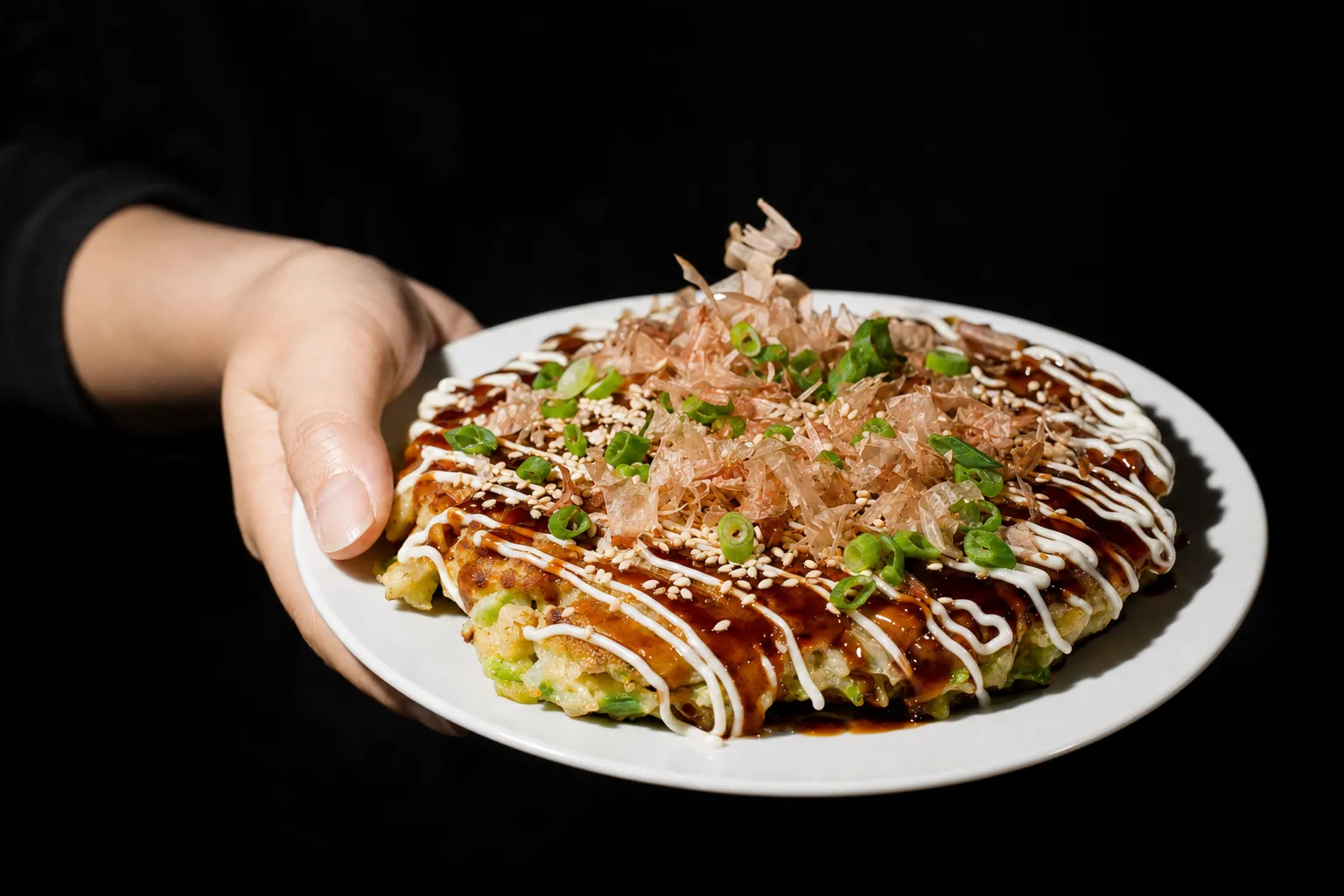 Ein Teller mit fertigem Okonomiyaki wird in die Kamera gehalten, mit Mayonnaise-Zickzack, Frühlingszwiebeln, Sesam und Bonito-Flakes.
