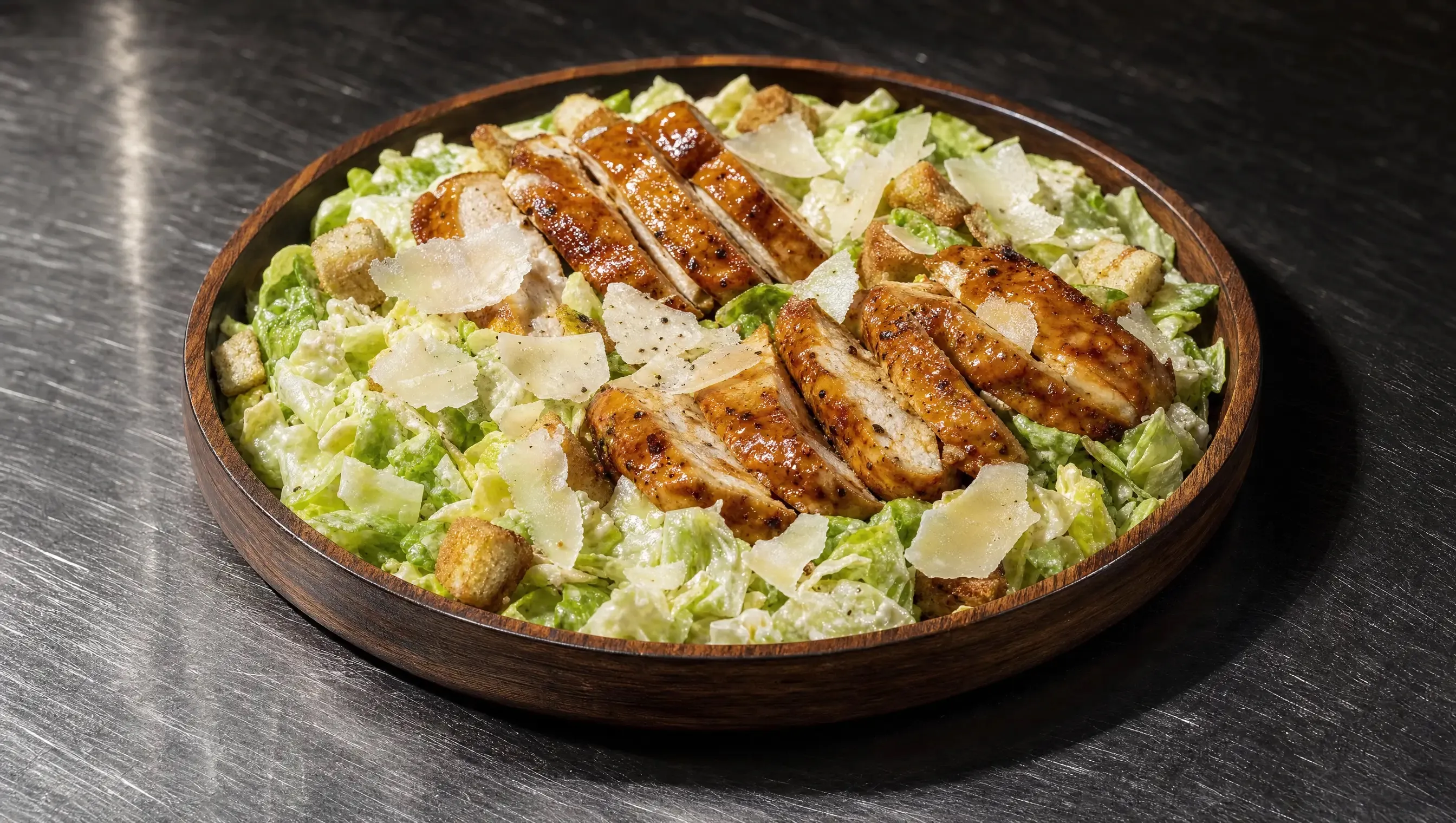 Dunkle Holzschale mit Caesar Salad, knackiger Blattsalat in cremigem Joghurt-Dressing, darauf goldbraun gebratene Hähnchenstreifen, gehobelter Parmesan und Croutons, mittig auf einer Edelstahl-Arbeitsplatte.