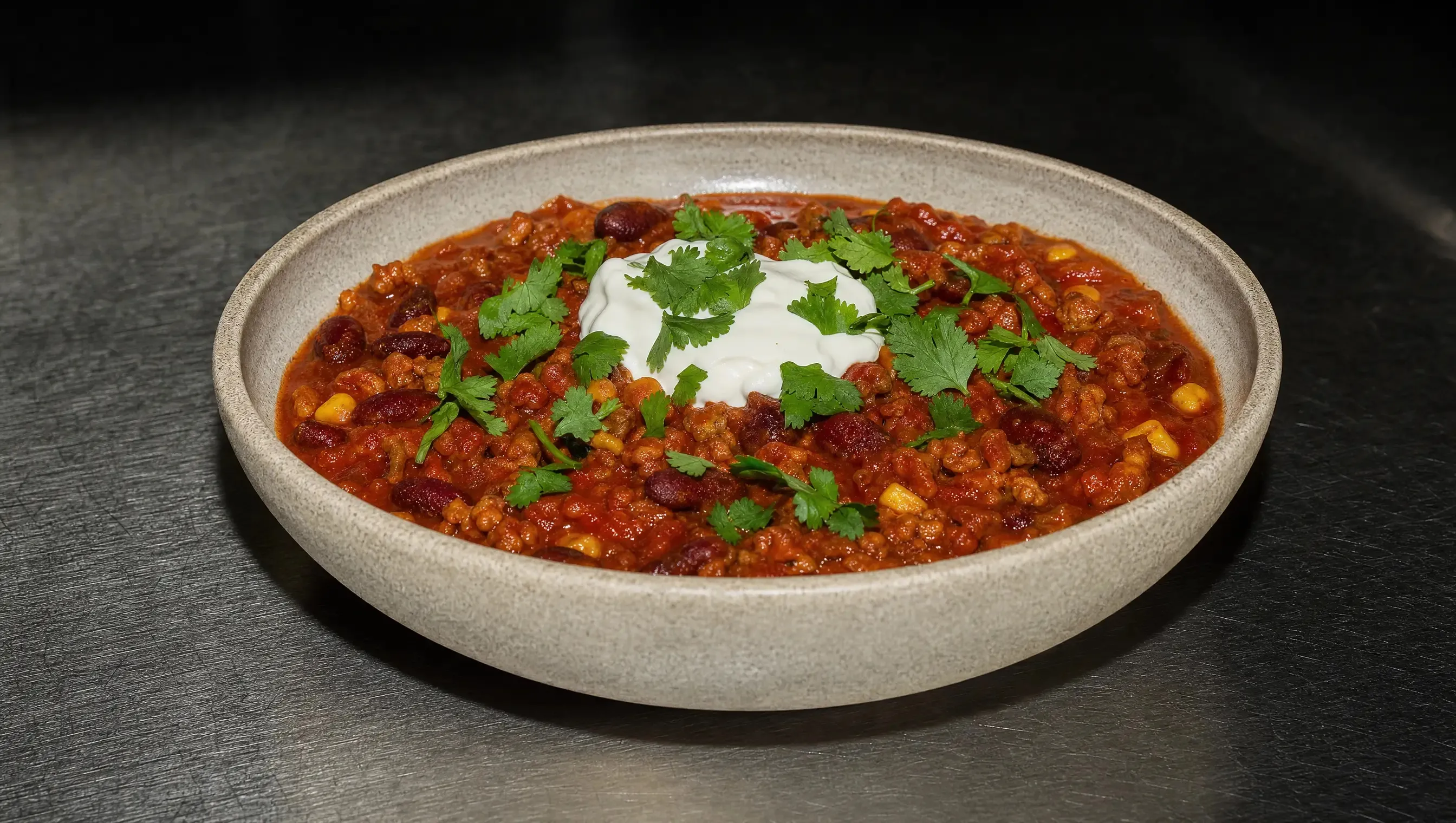 Eine helle Steingutschale mit Chili sin Carne aus Sojaschnetzel, Kidneybohnen und Mais in dicker Tomatensauce, in der Mitte ein Klecks Joghurt mit frischem Koriander, auf einer Edelstahl-Arbeitsfläche.