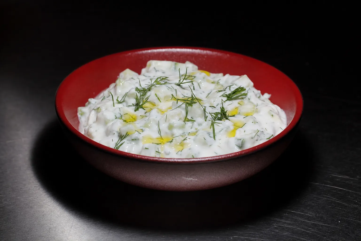 Rote Keramikschüssel mit cremigem Tzatziki, Gurkenstücken, frischem Dill und einem Schuss Olivenöl auf einer dunklen Fläche.