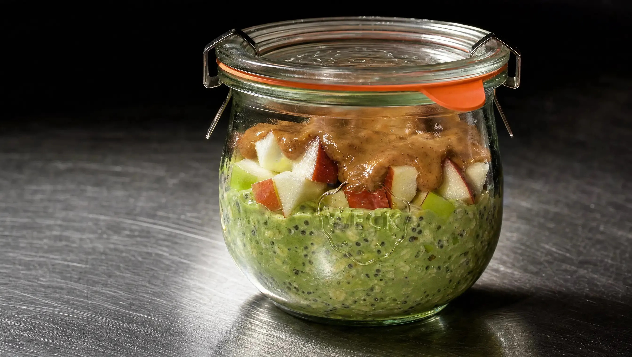 WECK-Glas mit grünen Matcha Overnight Oats, getoppt mit roten und grünen Apfelwürfeln und einer Haube Nussmus, mittig auf einer dunklen Edelstahl-Arbeitsplatte.
