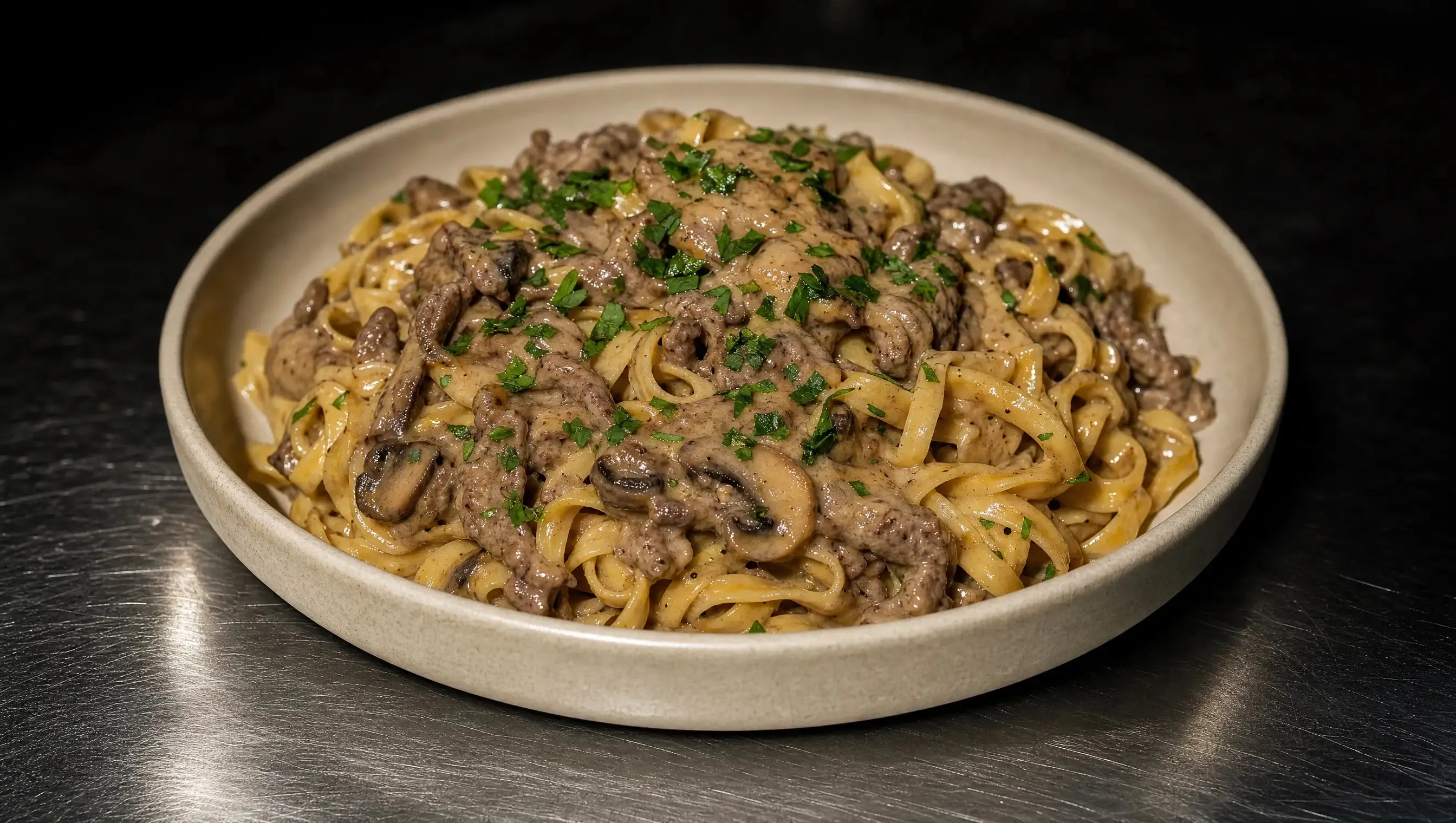 Cremeweiße Keramikschüssel mit Tagliatelle in cremiger Rinder-Pilz-Sauce, garniert mit gehackter Petersilie, mittig auf einer dunklen Edelstahl-Arbeitsplatte.
