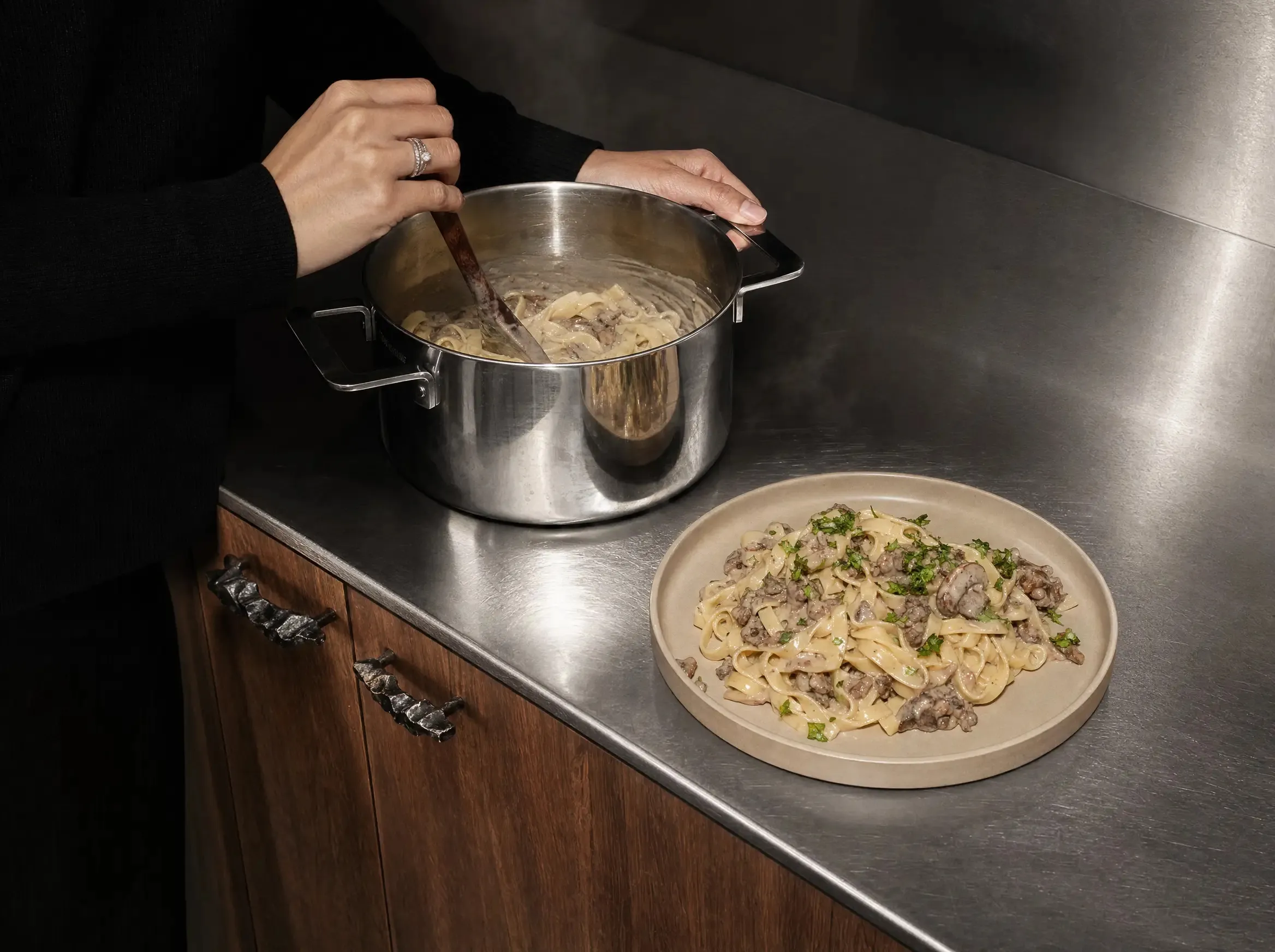 Hände rühren das Beef Stroganoff mit einem Löffel in einem großen Edelstahltopf um, daneben steht ein bereits angerichteter Teller mit Nudeln und Petersilie auf der Arbeitsplatte.