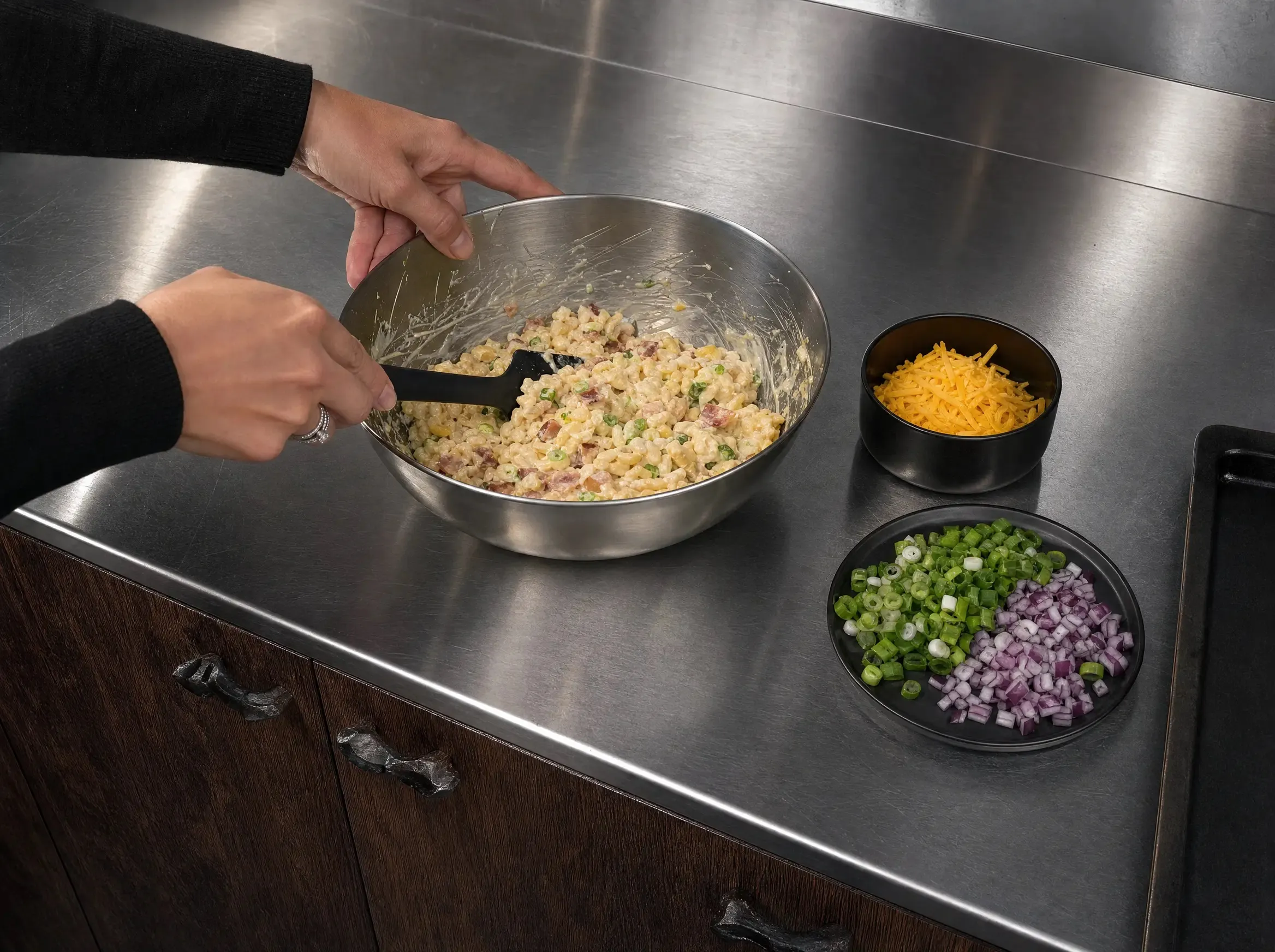 Hände vermengen Spätzle mit Frischkäsesauce, Frühlingszwiebeln und Speckwürfeln in einer Edelstahlschüssel, daneben Schälchen mit geriebenem Cheddar und geschnittenen Zwiebeln.