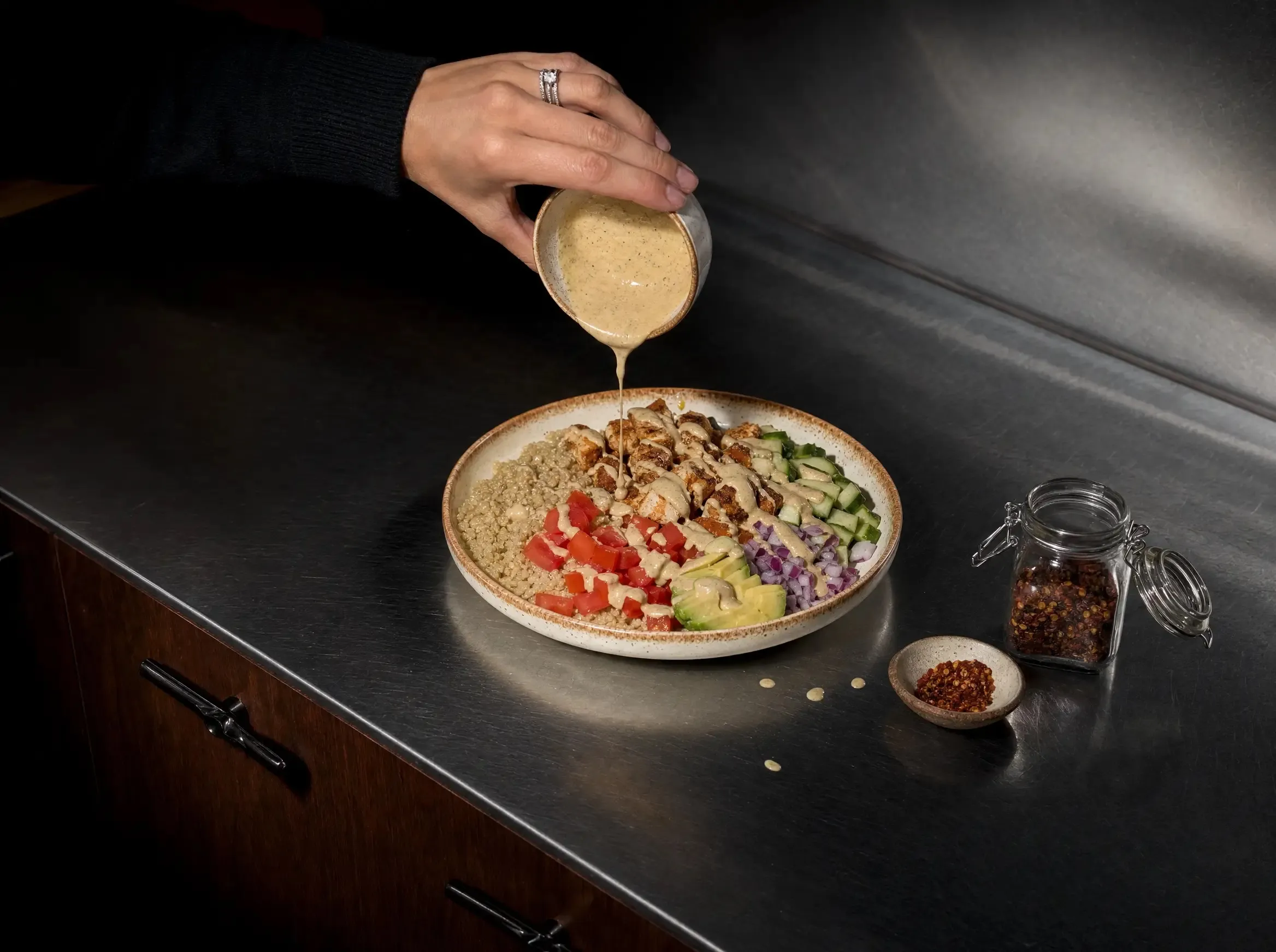 Eine Hand gießt cremiges Tahini-Dressing aus einem kleinen Schälchen über die angerichtete Quinoa Bowl, daneben ein Bügelglas und ein Schälchen mit Chiliflocken auf der Arbeitsfläche.