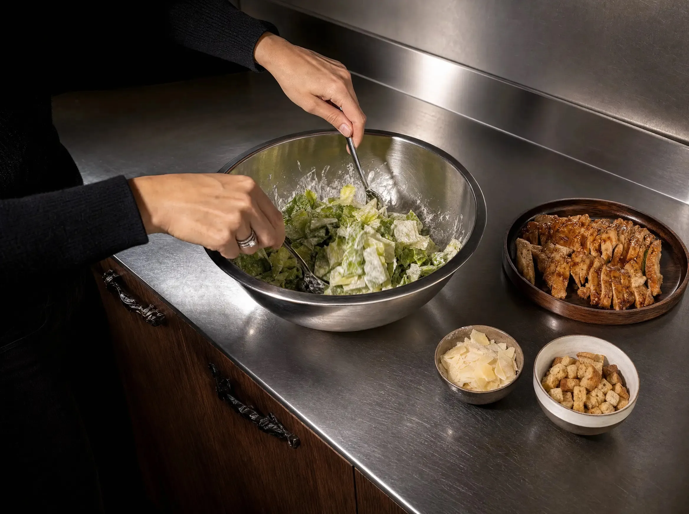 Hände vermischen geschnittenen Blattsalat mit dem Joghurt-Dressing in einer großen Edelstahlschüssel, daneben Schälchen mit Parmesan und Croutons sowie ein Brett mit aufgeschnittenem Hähnchen.