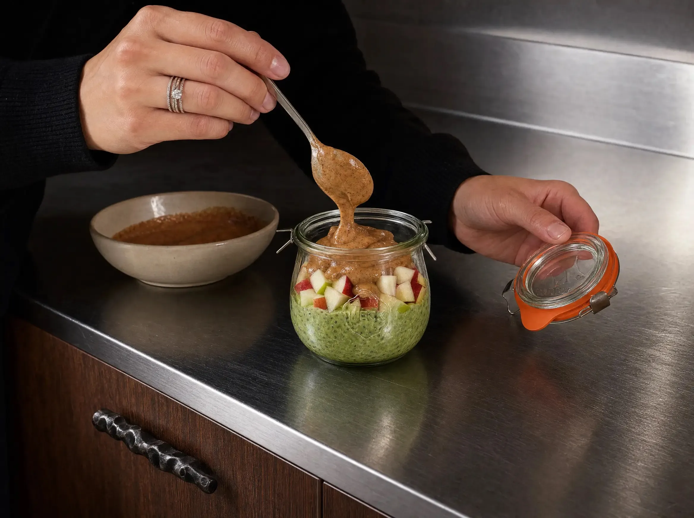 Eine Hand löffelt Nussmus auf die mit Apfelwürfeln belegten Matcha Overnight Oats im WECK-Glas, die andere Hand hält den abgenommenen Glasdeckel, daneben eine Schale mit Nussmus auf der Edelstahl-Arbeitsplatte.