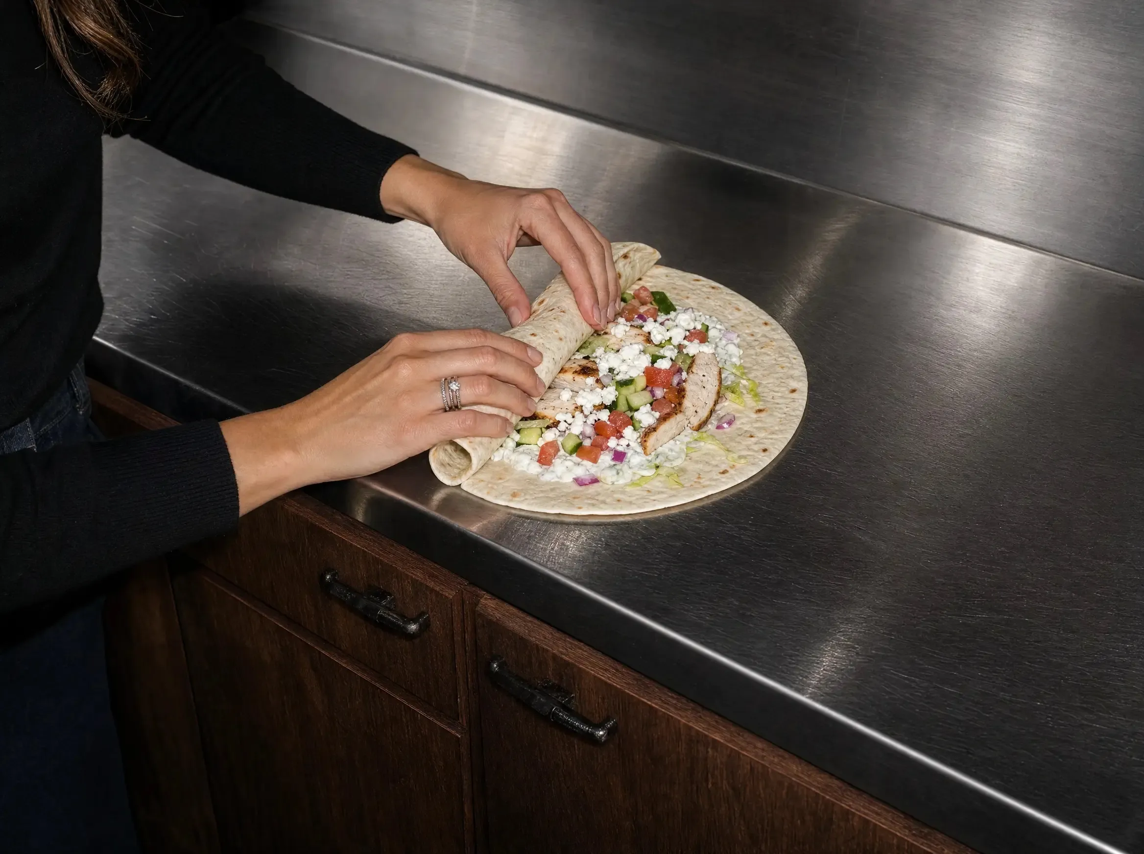 Zwei Hände rollen eine belegte Tortilla über der Füllung aus Hähnchen, Feta und Gemüse zusammen, auf einer Edelstahl-Arbeitsplatte.