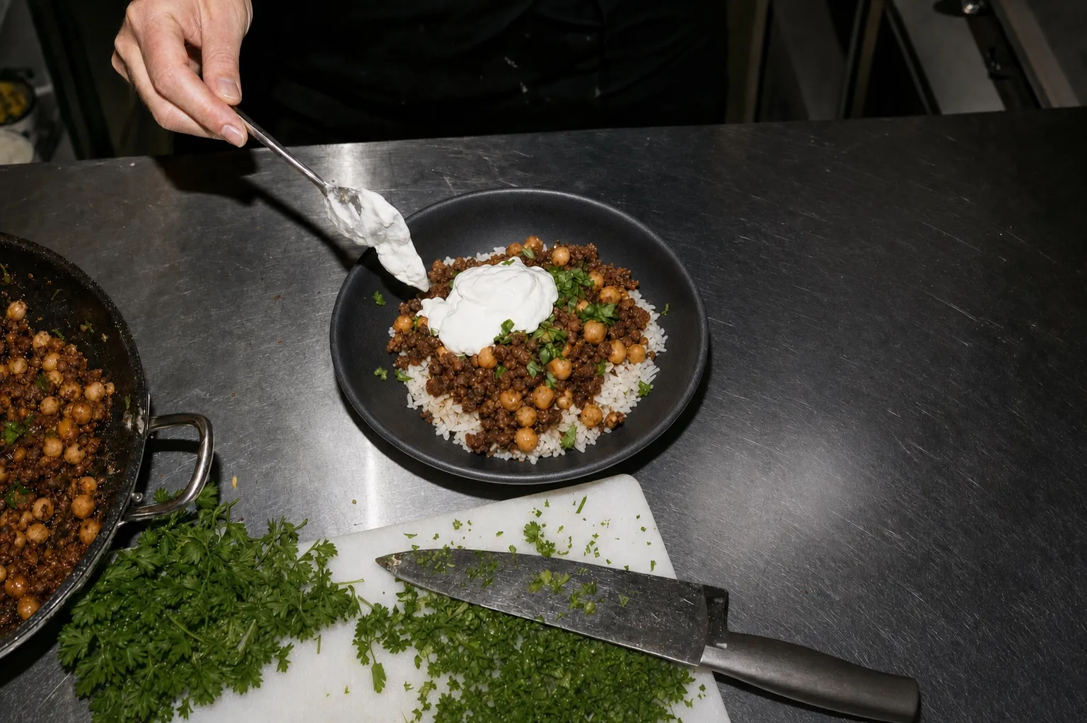 Frauenhände geben Joghurt-Topping über eine Schüssel Hackfleisch-Kichererbsen-Pfanne auf Reis, Edelstahlküche im Hintergrund.